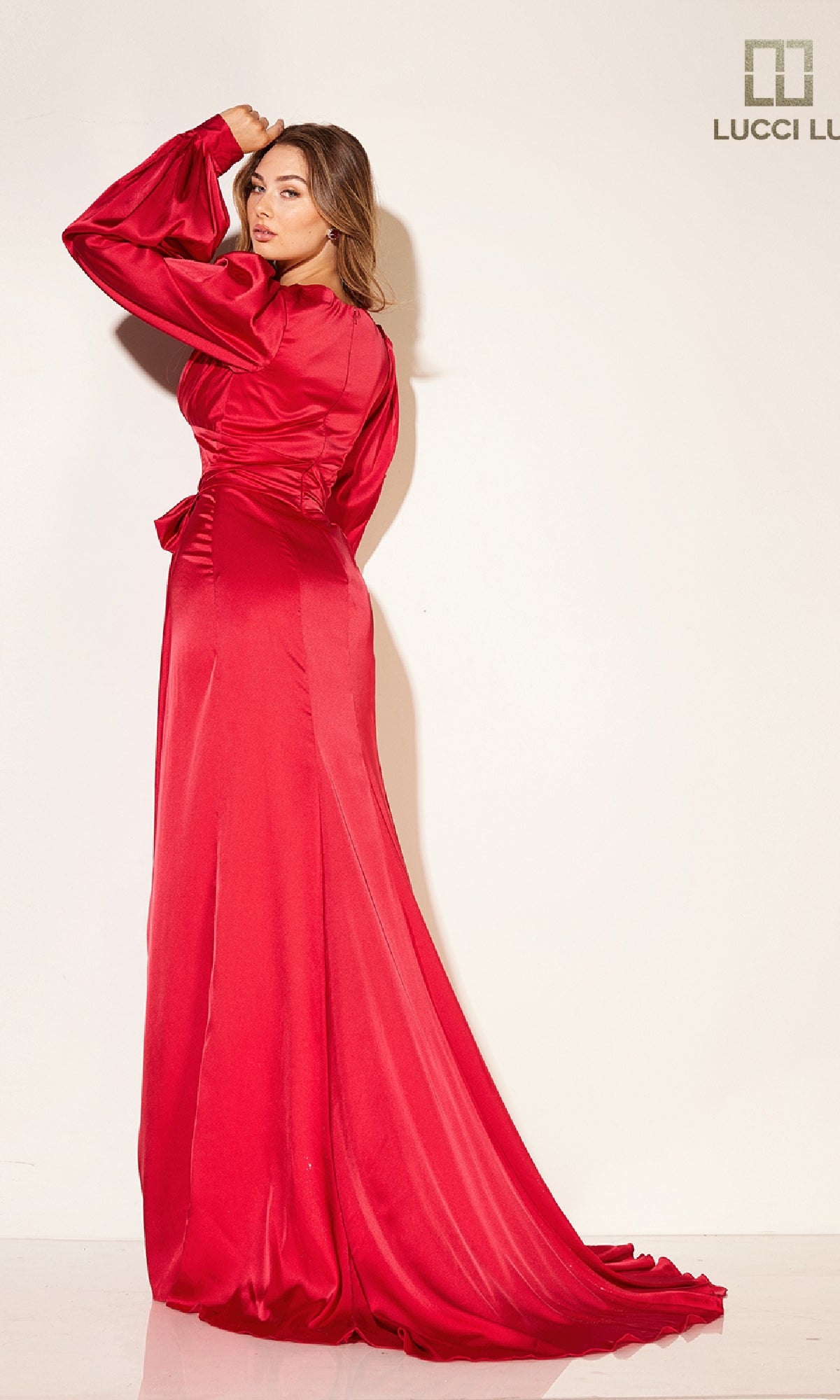 Woman wearing the Lucci Lu C8088 long formal dress with a wrap-style skirt and long sleeves, standing against a light background and looking over her shoulder.